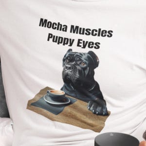 May include: White t-shirt with the text "Mocha Muscles Puppy Eyes" and a black dog illustration. The dog is shown with a coffee cup on a wooden tray. The dog has brown eyes.