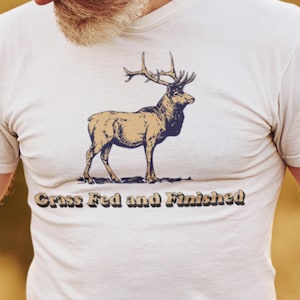 May include: A white t-shirt with a brown and blue illustration of an elk with large antlers. The text "Grass Fed and Finished" is printed in yellow below the elk.