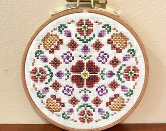 May include: A circular embroidery hoop with a white fabric background featuring a floral design in shades of red, green, purple, and gold. The design is symmetrical and features a central flower with smaller flowers and leaves surrounding it.