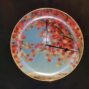 May include: A round wall clock featuring a light blue background with a detailed illustration of a tree with vibrant red and orange maple leaves. The clock has a thin gold-toned rim and two black hands positioned to indicate the time on the face.