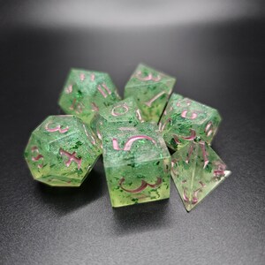 Puede incluir: Un conjunto de dados verdes y rosas con purpurina incrustada en la resina. Los dados son un d4, d6, d8, d10, d% y d20.