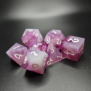 Puede incluir: Un conjunto de siete dados de color morado y blanco con números plateados. Los dados están hechos de un material translúcido, y los números son visibles a través del material.