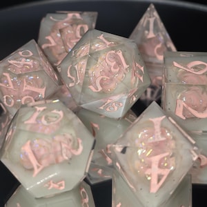 Puede incluir: Un conjunto de dados transparentes con números rosas y diseños florales en su interior. Los dados son multifacéticos y están sobre un fondo oscuro. Para juegos de mesa.