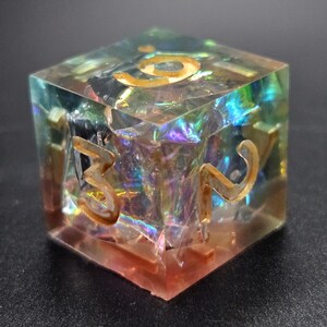 Puede incluir: Un cubo transparente iridiscente con números dorados 1, 2, 3, 4, 5 y 6 en cada lado. El cubo está hecho de resina y tiene un efecto brillante y holográfico.