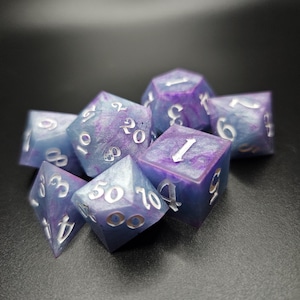 Puede incluir: Un conjunto de siete dados poliédricos en tonos azules y morados con números plateados. Los dados tienen un aspecto veteado y están colocados sobre una superficie oscura. Estos dados se utilizan para juegos de rol de mesa.