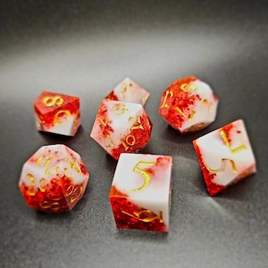 Puede incluir: Conjunto de seis dados blancos y rojos con números dorados. Los dados tienen un aspecto moteado y marmoleado.