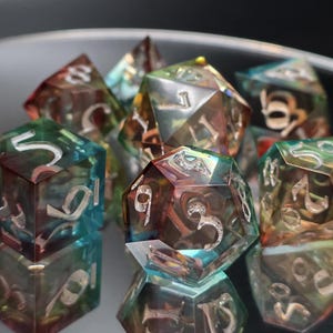 Puede incluir: Un conjunto de dados poliédricos iridiscentes con números plateados. Los dados tienen una apariencia colorida y translúcida, con tonos de verde, azul y rojo. Los dados están diseñados para juegos de mesa.