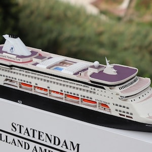 Pode incluir: Modelo de navio de cruzeiro com casco branco, fundo preto e detalhes roxos. O navio tem vários conveses, botes salva-vidas e uma chaminé. O nome "STATENDAM" é visível na lateral. O modelo está sobre uma superfície branca.