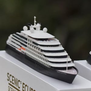 May include: A detailed miniature cruise ship model with a black hull and white decks. The model features multiple levels, windows, and a top deck with antennas. It is displayed on a white surface.