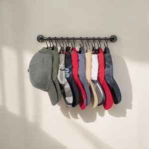 May include: A black metal hat rack mounted on a beige wall, displaying a collection of baseball caps. The caps are in various colors, including gray, red, and camouflage patterns. The rack has multiple hooks to hold the hats.