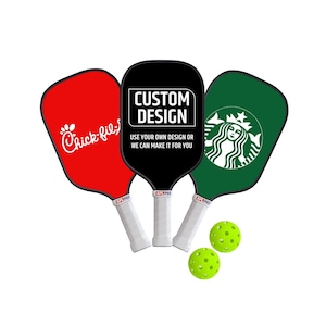 May include: Three pickleball paddles with white handles and APL PRO branding. The left paddle is red with a white logo, the center is black with white text reading CUSTOM DESIGN USE YOUR OWN DESIGN OR WE CAN MAKE IT FOR YOU, and the right is green with a white logo. Two neon yellow pickleballs are included.