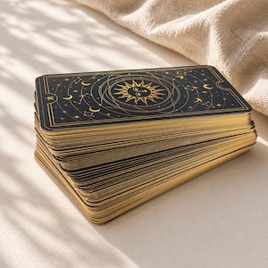 May include: A deck of rectangular tarot cards with black backs featuring a gold foil illustration of a sun with a face, surrounded by orbital lines, stars, and crescent moons. The edges of the cards are finished with a metallic gold gilded border.