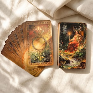 May include: A deck of tarot cards with a fan of cards showing a circular vine design on the back and one card featuring an illustration of a figure with antlers holding a staff and shield, labeled THE EMPRESS at the bottom in gold text.