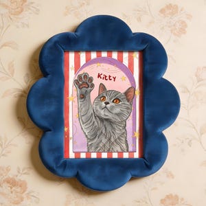 May include: A blue velvet framed picture of a cat. The artwork shows a gray cat raising a paw, with the name "Kitty" above. The background has red and white stripes and a purple arch.