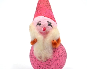 May include: A small gnome ornament featuring a bright pink glittered conical hat and a matching round pink glittered base. The figure has a pale pink face, black painted eyes, a small red nose, and a fluffy white beard with orange arms.