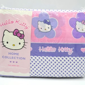 May include: A Hello Kitty Home Collection full sheet set in plastic packaging. The package features a pink and purple design with Hello Kitty character graphics, flower shapes, and polka dots. The text reads Hello Kitty Home Collection.