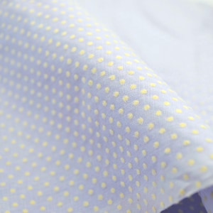 May include: Close-up of a light blue fabric with a pattern of small, evenly spaced yellow dots. The fabric is folded, showing texture and depth. The material appears to be a lightweight cotton or linen.