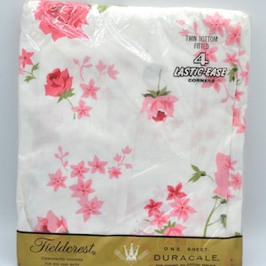 May include: A packaged twin-size fitted sheet with a floral pattern. The white sheet features pink roses and smaller pink flowers with green leaves. The packaging reads "Fieldcrest" and "DURACALE."