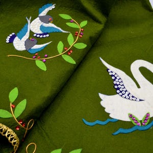 May include: An olive green fabric featuring embroidered and appliquéd designs of a white swan on blue water and two smaller birds perched on a branch. The fabric is decorated with small pink and red sequins and has a gold metallic lace trim edge.