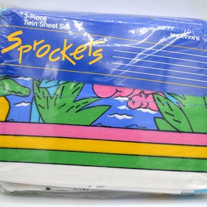 May include: A 3-piece twin sheet set in plastic packaging labeled Sprockets and Mervyn's. The fabric features a colorful graphic print of green crocodiles, pink hippos, and tropical foliage against a blue background, with horizontal stripes.