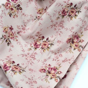May include: A light beige fabric featuring a repeating floral pattern of deep pink and muted yellow roses with dark green leaves and thin, winding brown stems. The textile has a soft, draped appearance with visible folds and a matte finish.