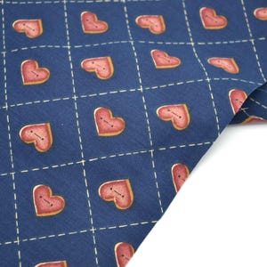 May include: A navy blue fabric with a repeating pattern of pink heart-shaped buttons. The buttons are outlined in gold and have two small black dots. The fabric is divided into squares by white dashed lines.