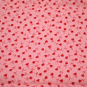 May include: Close-up of a pink fabric with a repeating pattern of small red hearts on stems. The hearts are evenly spaced across the textured surface, creating a romantic and delicate design. Ideal for crafting or sewing projects.
