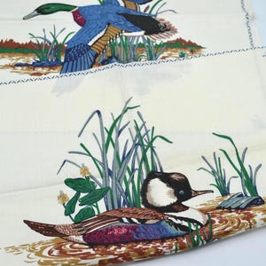 May include: Two off-white fabric napkins with duck illustrations. One shows a duck in flight with blue, brown, and green details. The other depicts a duck resting in water with similar colors and details.