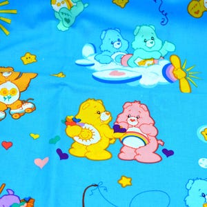 May include: Light blue fabric featuring colorful Care Bears characters. The design includes yellow, pink, and blue bears, along with rainbows, stars, and hearts. Some bears are on roller skates or flying in a plane.