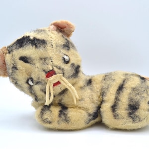 May include: A vintage plush toy cat with black and white stripes, a pink nose, and white yarn whiskers. The cat is lying on its side with its head turned to the left.