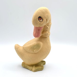 May include: A vintage, light yellow duck figurine with a pink beak and orange eyes. The duck has a textured surface and is standing on a green base. The figurine is a charming piece of decor.