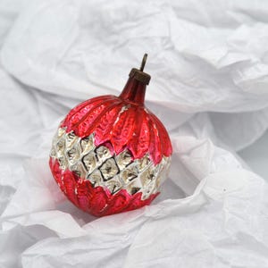 May include: A vintage Christmas ornament with a red and silver design. The top of the ornament is red with a textured pattern, while the bottom half features a silver, diamond-shaped pattern. The ornament has a metal cap and loop for hanging.