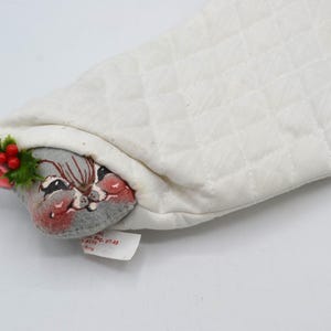 May include: A white quilted fabric Christmas stocking featuring a gray mouse head peeking out from the top. The mouse has pink ears, red berries, and green holly leaves attached. A green and red floral pattern is on the upper right side.