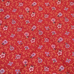 May include: Red fabric with a repeating floral pattern. The design features small white, pink, and blue flowers with green leaves and gold vines. The fabric is suitable for quilting and crafting projects.