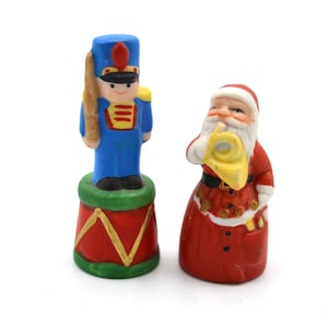 May include: Two small Christmas figurines. One is a blue toy soldier with a red and gold drum. The other is Santa Claus in a red suit, holding a yellow horn. Both are painted with bright colors.