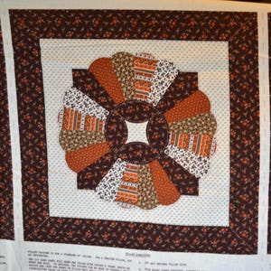 May include: Close-up of a fabric square featuring a circular, fan-like design in shades of brown, orange, and white. The central design is surrounded by a white background with small gray dots, framed by a dark brown border with floral patterns. The overall theme is quilting.