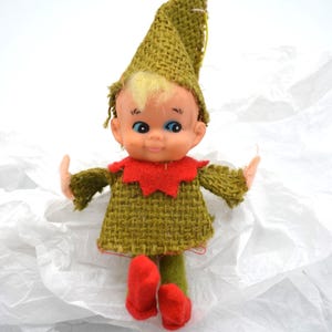May include: A vintage elf doll with a green burlap outfit, red collar and shoes, and a matching hat. The doll has a smiling face with blue eyes and blonde hair. The doll is standing on a white surface.
