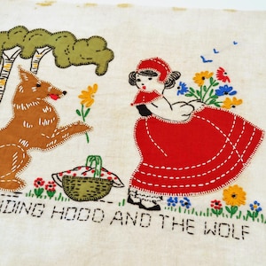 May include: A rectangular beige fabric panel featuring colorful embroidery of a girl in a red hood and a brown wolf. The scene includes a green tree, a picnic basket, and flowers. The text RED RIDING HOOD AND THE WOLF is stitched in black.