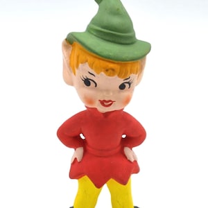 May include: A small figurine of an elf with a green pointed hat, orange hair, and large ears. The elf wears a red tunic with a scalloped hem, yellow pants, and long, pointed black shoes. The face features painted black eyes, red lips, and rosy cheeks.