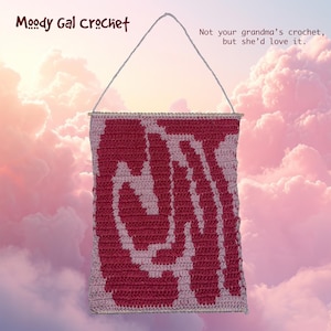 Peut inclure: Une tenture murale au crochet faite à la main avec un motif abstrait rose et rouge. La pièce est rectangulaire, avec un fond rose clair et un motif rouge vif. Le texte "Moody Gal Crochet" et "Not your grandma's crochet, but she'd love it" sont également visibles.