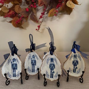May include: Four white and blue ceramic oyster shell ornaments with a gold trim, each featuring a blue nutcracker soldier design. The ornaments hang from navy blue ribbons and are displayed on black metal stands.
