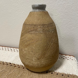 May include: A brown ceramic vase with a white rim and a textured surface. The vase has a rounded shape and is decorated with lines.