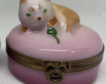 May include: A small, pink porcelain trinket box with a white cat figurine on top. The cat is holding a green ball in its paw.