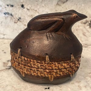 May include: A handcrafted, dark brown wooden container with a frog-shaped lid. The container's base features a woven, light brown band. The frog lid is intricately carved, adding a unique touch to this decorative piece.