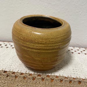 May include: A small, round, ceramic planter with a light brown glaze. The planter has a slightly textured surface and a wide, round opening.