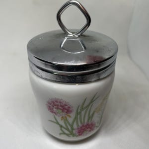 May include: White ceramic egg cup with a silver-colored metal lid and handle. The cup is decorated with pink and yellow floral designs and green leaves. The handle is a square shape.