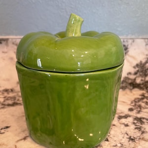 May include: A ceramic, green bell pepper-shaped container with a lid. The container is a vibrant shade of green, with a glossy finish. The lid is shaped like the top of a bell pepper, complete with a stem. The container is likely used for storage.