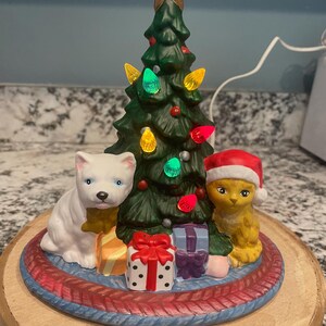 May include: A ceramic Christmas tree with colorful lights, a gold star, and two animal figurines. A white dog with a yellow scarf sits next to a yellow cat wearing a Santa hat. Presents are at the base of the tree.