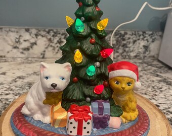 Puede incluir: Un árbol de Navidad de cerámica con luces de colores, una estrella dorada y dos figuras de animales. Un perro blanco con una bufanda amarilla se sienta junto a un gato amarillo con un gorro de Papá Noel. Los regalos están en la base del árbol.