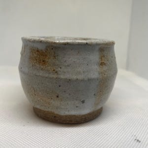 May include: A small, handmade ceramic cup with a rounded shape. The cup has a light gray exterior with brown and tan accents. The base is a darker brown color. The cup appears to be unglazed and has a rustic, textured finish.